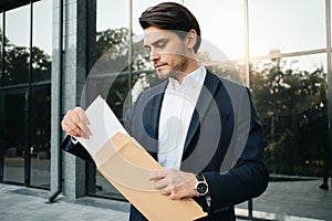 Young handsome bearded brunette man in white shirt and classic jacket standing dreamily opening envelope with glass