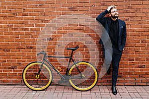 Young handsome bearded business man in suit and glasses with hands on head standing near his bicycle on brick wall background