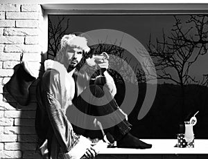 Young handsome bearded christmas man in red santa claus hat and coat with muscular bare torso sitting on windowsill