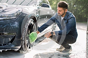 Young handsome bearded guy holding green sponge, washing car wheel with foam. Cleaning of modern rims of luxury blue