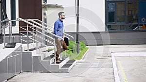 Young handsome bearded man in blue shirt and brown trousers is going outside. Student walking outdoors happily after