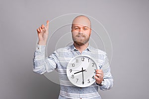 Young handsome bearded man holding clock and pointing up at copyspace