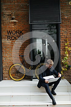 Young handsome bearded man sitting on the street stairs working on laptop near his bicycle