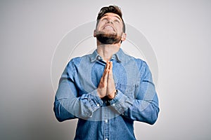 Young handsome blond man with beard and blue eyes wearing casual denim shirt begging and praying with hands together with hope