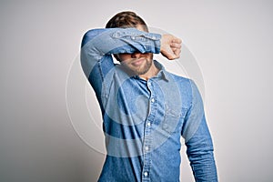 Young handsome blond man with beard and blue eyes wearing casual denim shirt covering eyes with arm, looking serious and sad