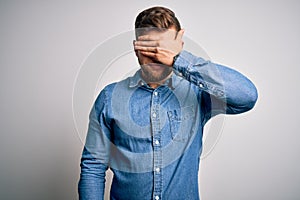 Young handsome blond man with beard and blue eyes wearing casual denim shirt covering eyes with hand, looking serious and sad