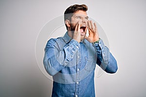 Young handsome blond man with beard and blue eyes wearing casual denim shirt Shouting angry out loud with hands over mouth