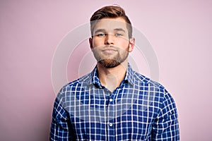 Young handsome blond man with beard and blue eyes wearing casual shirt standing with serious expression on face