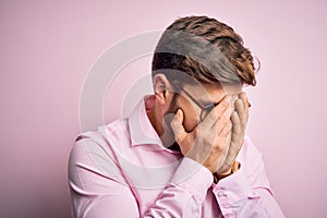 Young handsome blond man with beard and blue eyes wearing pink shirt and glasses with sad expression covering face with hands