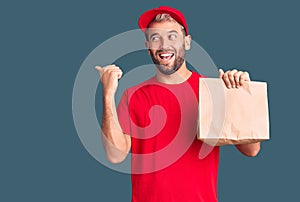 Young handsome blond man holding delivery paper bag pointing thumb up to the side smiling happy with open mouth