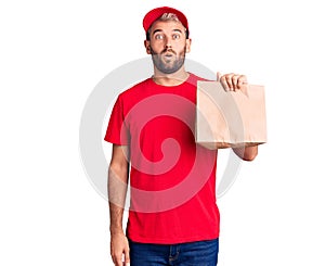 Young handsome blond man holding delivery paper bag scared and amazed with open mouth for surprise, disbelief face