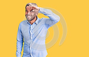 Young handsome blond man wearing elegant shirt very happy and smiling looking far away with hand over head