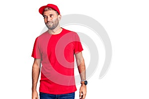 Young handsome blond man wearing t-shirt and cap looking away to side with smile on face, natural expression