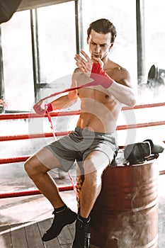 Young handsome boxer pulls bandage before the fight or training sitting on the red barrel in boxing ring. Boxing Fighter