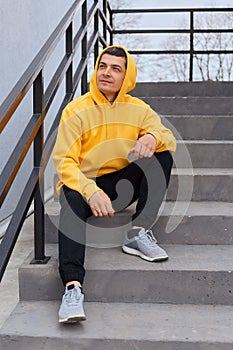 Young handsome brunette man sitting on stairs outdoors, looking away with pensive dreaming expression, thinks about something