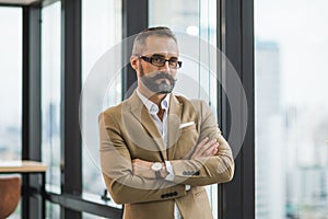 Young handsome business bearded man standing near window with arms crossed in office work place