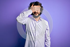 Young handsome business man with beard wearing shirt standing over purple background covering eyes with hand, looking serious and