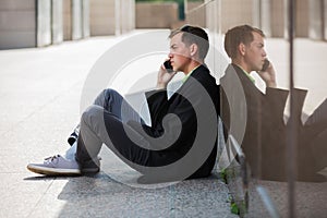 Young handsome business man calling on cell phone sitting on sidewalk