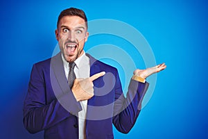 Young handsome business man over blue isolated background amazed and smiling to the camera while presenting with hand and pointing