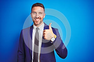 Young handsome business man over blue isolated background doing happy thumbs up gesture with hand