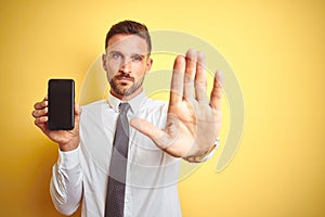 Young handsome business man showing smartphone screen over yellow isolated background with open hand doing stop sign with serious