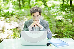 Young handsome business man thinking over laptop at office table in green park. Business concept.