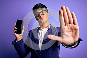 Young handsome business man wearing jacket and glasses holding smartphone showing screen with open hand doing stop sign with
