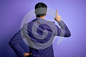 Young handsome business man wearing jacket and glasses over isolated purple background Posing backwards pointing ahead with finger