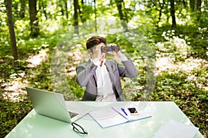 Young handsome business man at work table office with laptop in green forest with binocular looking for competitors. Business conc