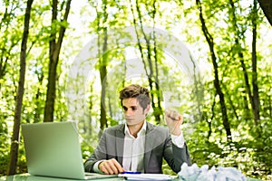 Young handsome business man at work table office with laptop in green forest with crumpled papers feeling exhausted. Business conc