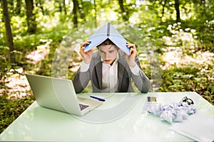 Young handsome business man at work table office with laptop in green forest with notebook over head, too much work. Business conc