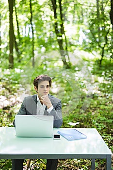 Young handsome business man working at laptop at office table with hand on chin thinking new idea in green park. Business concept.