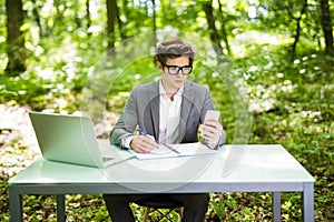 Young handsome business man working at laptop at office table use phone and texting in green park. Business concept.