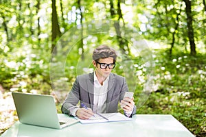 Young handsome business man working at laptop at office table use phone and texting in green park. Business concept.