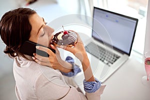 A young handsome business woman is enjoying while eating a delicious donut with a pleasure and talking on a phone in a pastry shop