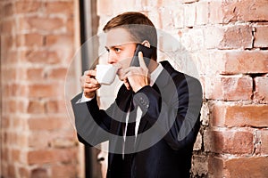 Young handsome businessman in black suit is drinking coffee and calling by the phone near brick wall