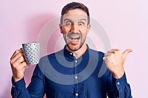 Young handsome businessman drinking mug of coffee over isolated pink background pointing thumb up to the side smiling happy with