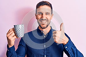 Young handsome businessman drinking mug of coffee over isolated pink background smiling happy and positive, thumb up doing