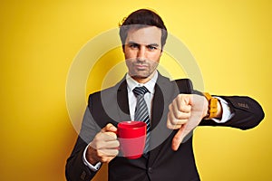 Young handsome businessman drinking red cup of coffee over isolated yellow background with angry face, negative sign showing