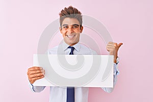 Young handsome businessman holding banner standing over isolated pink background pointing and showing with thumb up to the side