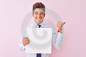 Young handsome businessman holding banner standing over isolated pink background pointing and showing with thumb up to the side