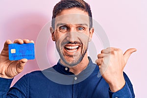 Young handsome businessman holding credit card over isolated pink background pointing thumb up to the side smiling happy with open