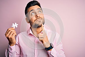 Young handsome businessman holding piece of puzzle over isolated pink background serious face thinking about question, very