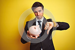 Young handsome businessman holding piggy bank over  yellow background with angry face, negative sign showing dislike with