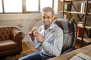 Young handsome businessman sit on chair and have lunch time in his own office. He hold burger and smile to camera