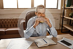 Young handsome businessman sit at table and biting burger in his own office. He has lunch time. Hungry young man devour