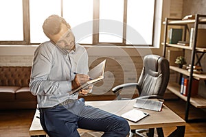 Young handsome businessman sit on table and talk on phone in his own office. He write in notebook. Business call