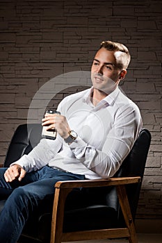 Young handsome businessman sitting on leather couch with phone in hand and thinking