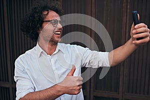 Young handsome businessman taking self portrait with his smart phone outdoors. Happy smiling male wearing white shirt and round