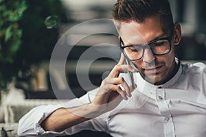 Young handsome businessman is talking on phone, sitting at table in cafe. Man in glasses makes call. Business person in